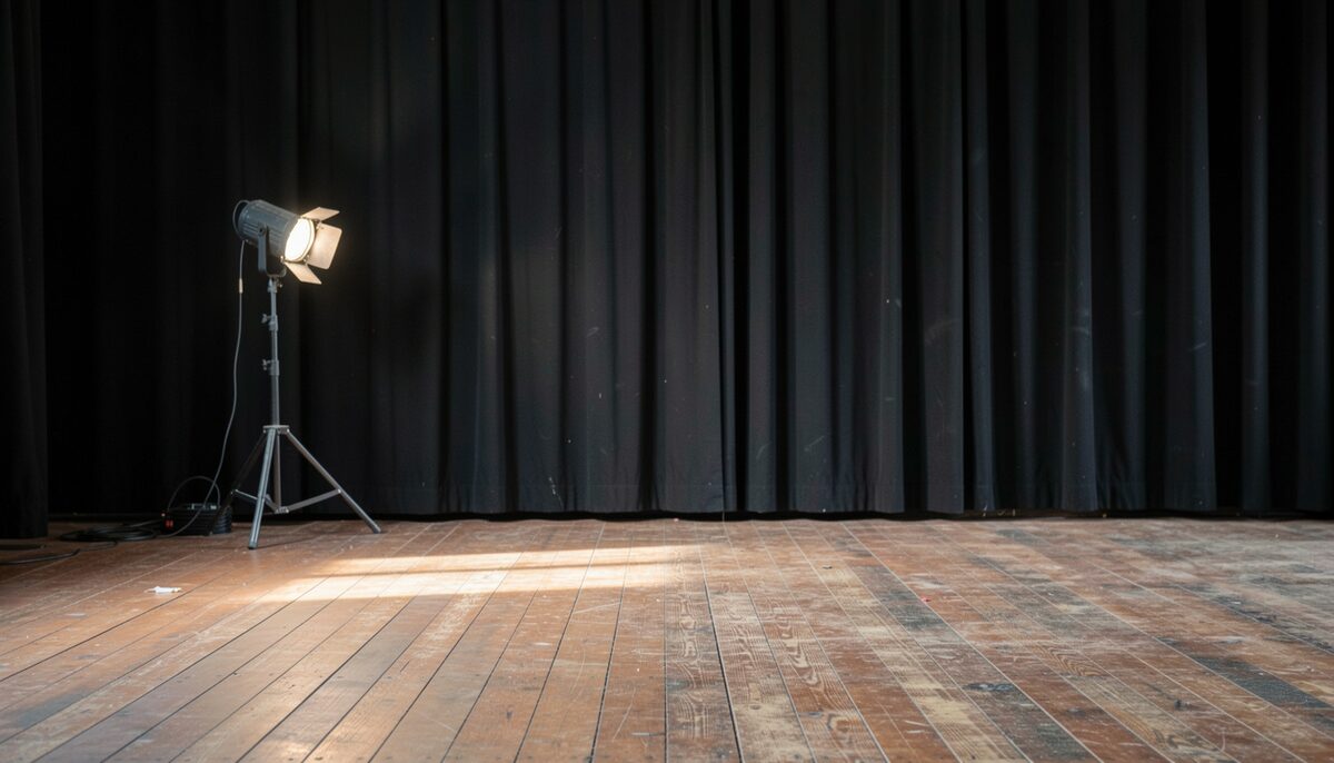The view from the wings of a stage: worn wooden floorboards, a hanging black curtain and a single work light on a stand. No paper, no poster, no sign, no screen, no label and no printed surface in the frame; no lettering, no words and no numbers anywhere.